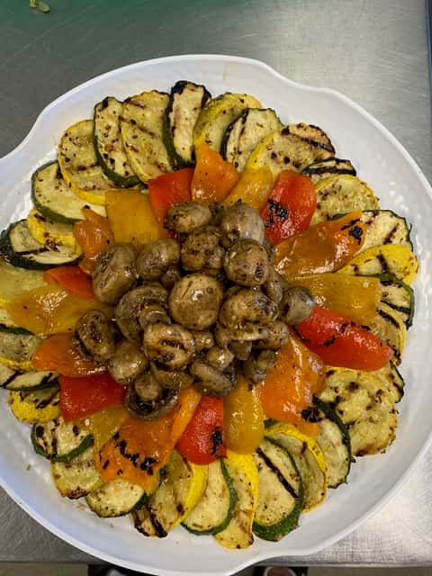 Grilled vegetable medley beautifully arranged on a white platter, featuring charred zucchini and yellow squash slices fanned around the edge, with vibrant roasted red and yellow bell peppers forming a ring, and a savory center of sautéed mushrooms.