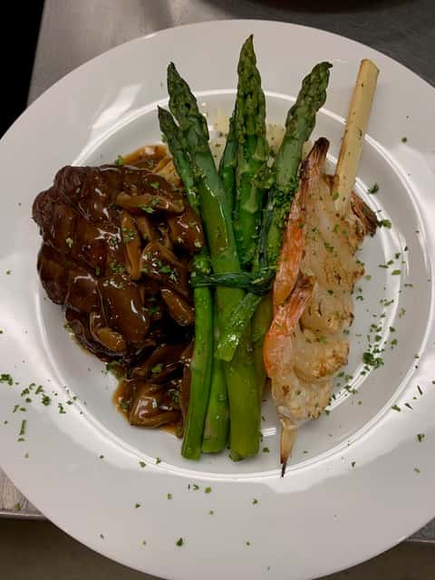 Surf and turf dinner plate featuring a grilled lobster tail skewer, tender steak topped with a savory mushroom demi-glace, and a bundle of steamed asparagus spears, garnished with fresh chopped herbs on a white plate.