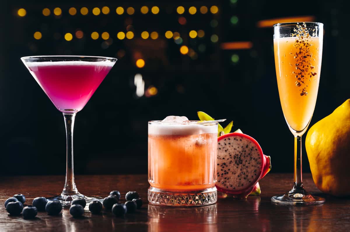 Three brightly colored New American craft cocktails on a dark wooden bar, including a magenta martini, a frothy orange old-fashioned glass drink, and an effervescent yellow champagne flute, garnished with blueberries and dragon fruit
