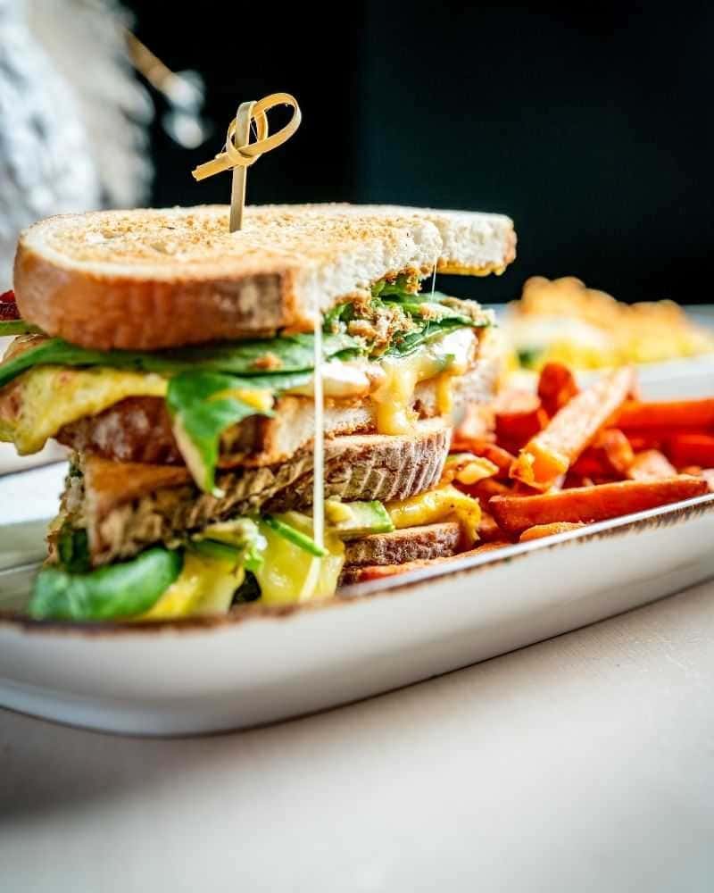 Gourmet grilled sandwich stacked with melted cheese, fresh greens, and sliced vegetables on toasted multigrain bread, served with crispy sweet potato fries on a modern restaurant plate with a warm, casual dining ambiance.