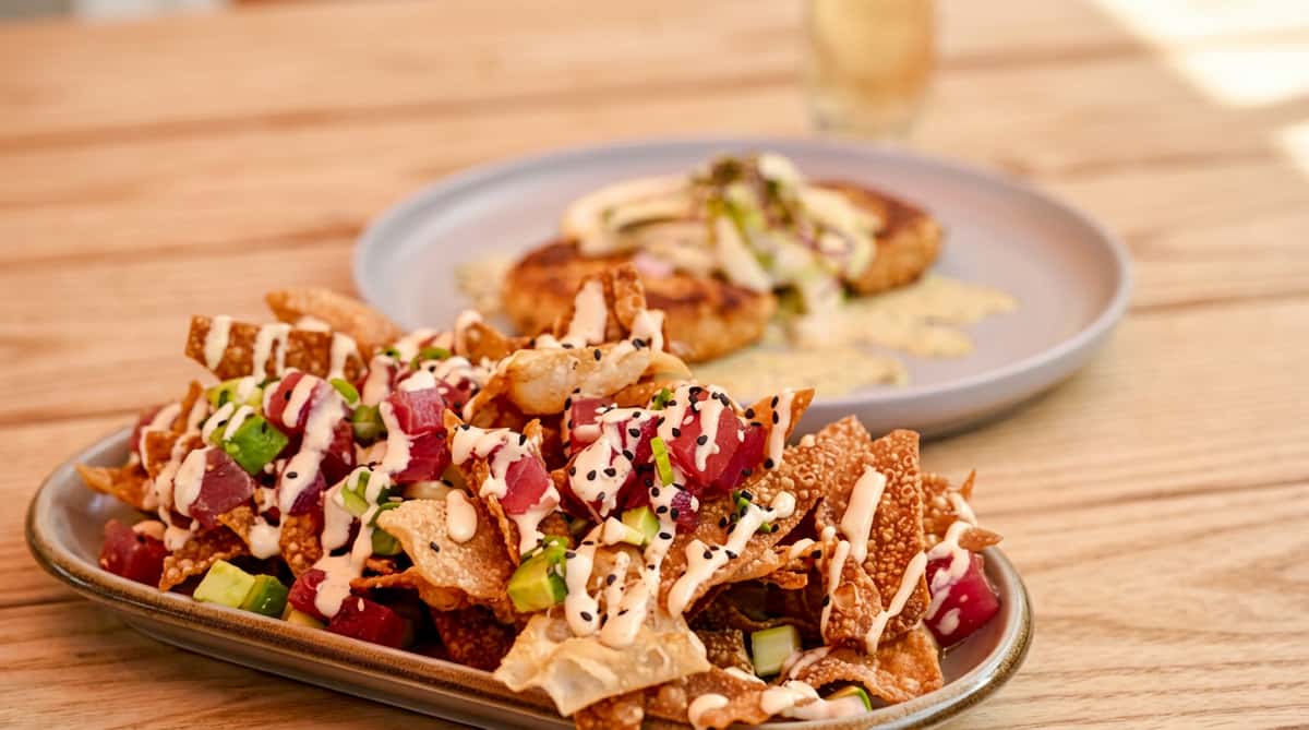 Tuna poke nachos with fried wonton chips, diced tuna, avocado, creamy sauce, and sesame seeds; blurred crab cakes in background.