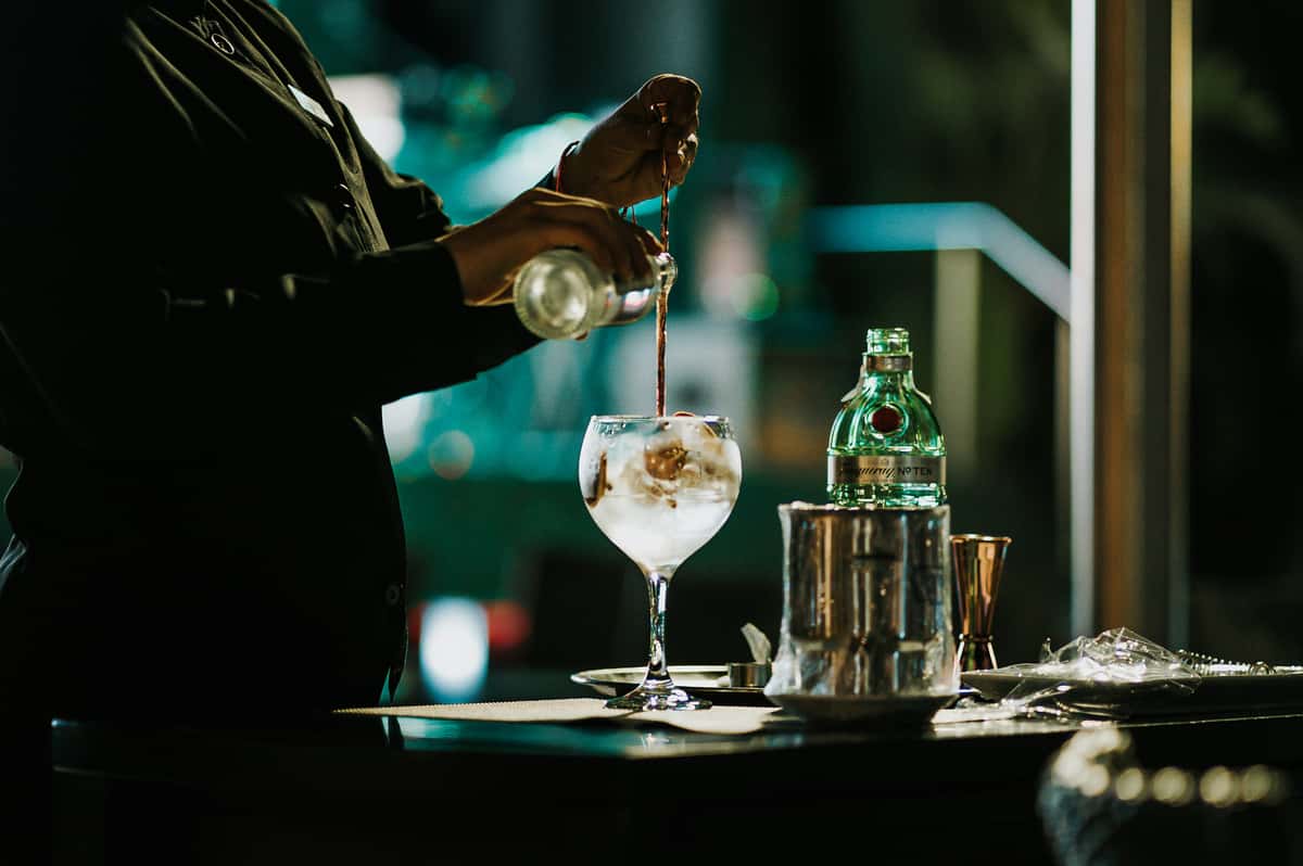 Bartender pouring tonic water into a gin cocktail over ice in a stemmed glass, with Fever-Tree bottle and bar tools nearby.