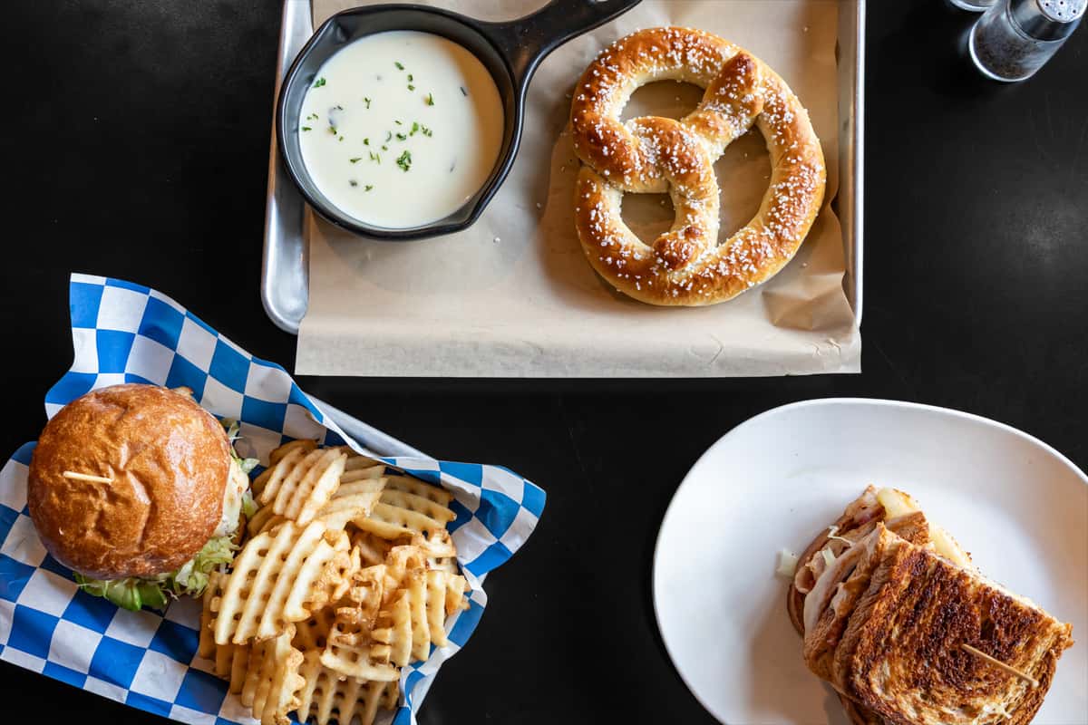 Flat-lay of classic bar fare including a soft salted pretzel with a skillet of creamy cheese dip, a grilled sandwich stacked with deli meats and melted cheese, and a burger with lettuce served alongside golden waffle fries on a blue-checkered liner.