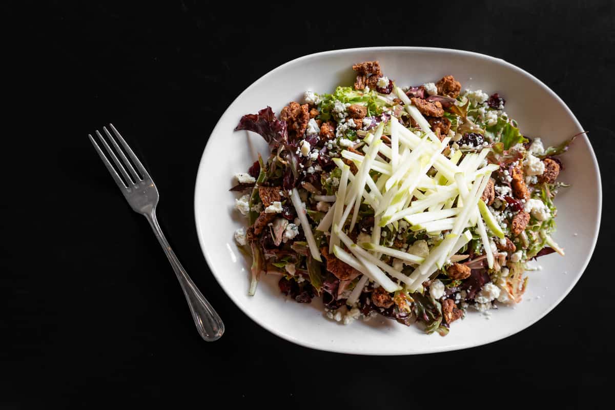 Overhead shot of a fresh mixed greens salad topped with julienned green apples, crumbled blue cheese, dried cranberries, candied pecans, and crunchy croutons, served on a white oval plate with a fork on a dark tabletop.