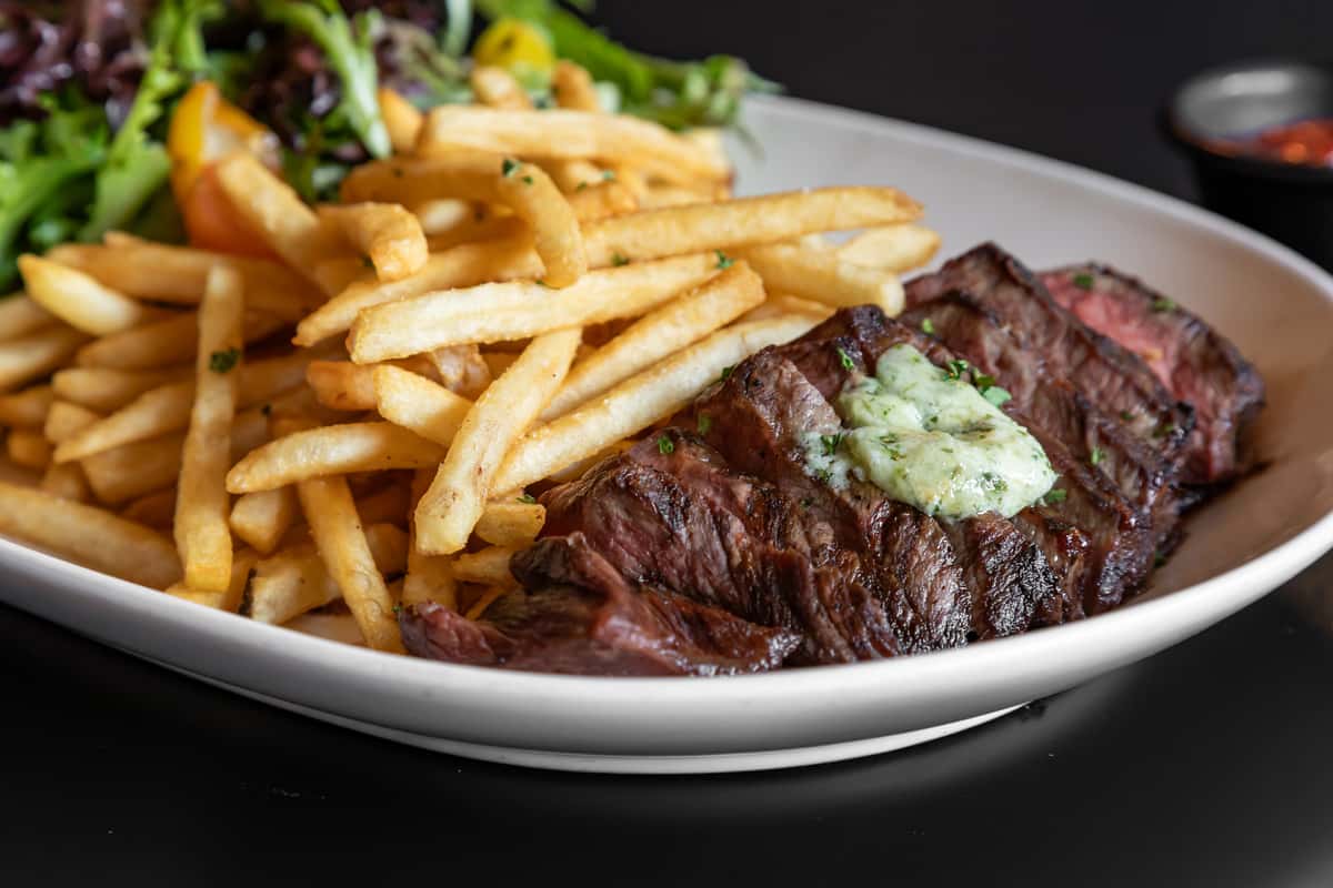 Close-up of a juicy grilled steak topped with herb butter, served with crispy golden fries and a fresh mixed green salad on a white plate at Rookies Sports Bar and Grill.