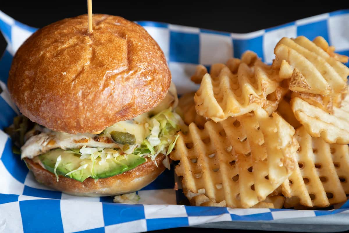 Close-up of a grilled chicken sandwich topped with shredded lettuce, melted cheese, jalapeños, and sliced avocado on a toasted brioche bun, served with a side of crispy waffle fries on a blue-checkered liner.