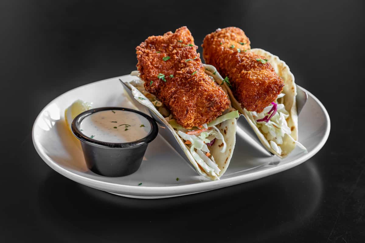 Two crispy fried fish tacos filled with shredded cabbage in flour tortillas, served with a side of tartar sauce.
