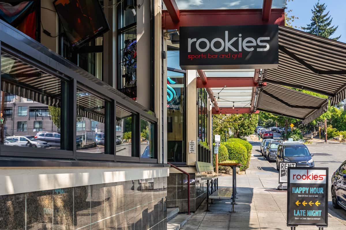 Close-up angle of the Rookies Sports Bar and Grill exterior, featuring floor-to-ceiling windows, flat-screen TVs visible inside, and a shaded sidewalk with a promotional sign for happy hour and late-night specials.