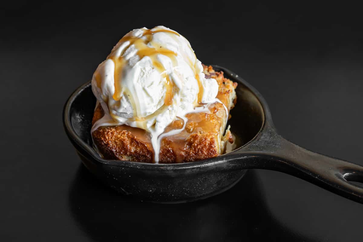 Warm bread pudding served in a cast-iron skillet, topped with a generous scoop of vanilla ice cream and rich caramel sauce.