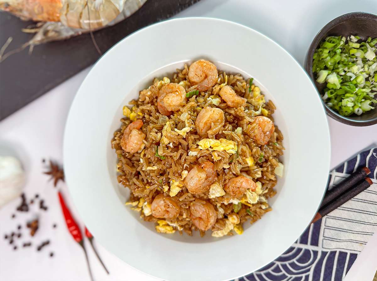 Bowl of shrimp fried rice with pieces of scrambled egg and green onions, garnished with shrimp on top, served on a white plate. In the background, there are whole spices, dried red chilies, and a small bowl of chopped green onions.