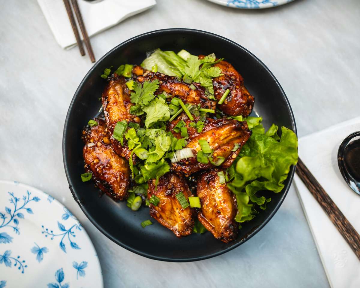 A black bowl filled with glazed chicken wings garnished with fresh cilantro and green onions, resting on a bed of lettuce. In the background, a decorative blue and white plate and chopsticks are visible, suggesting an Asian dining setting.