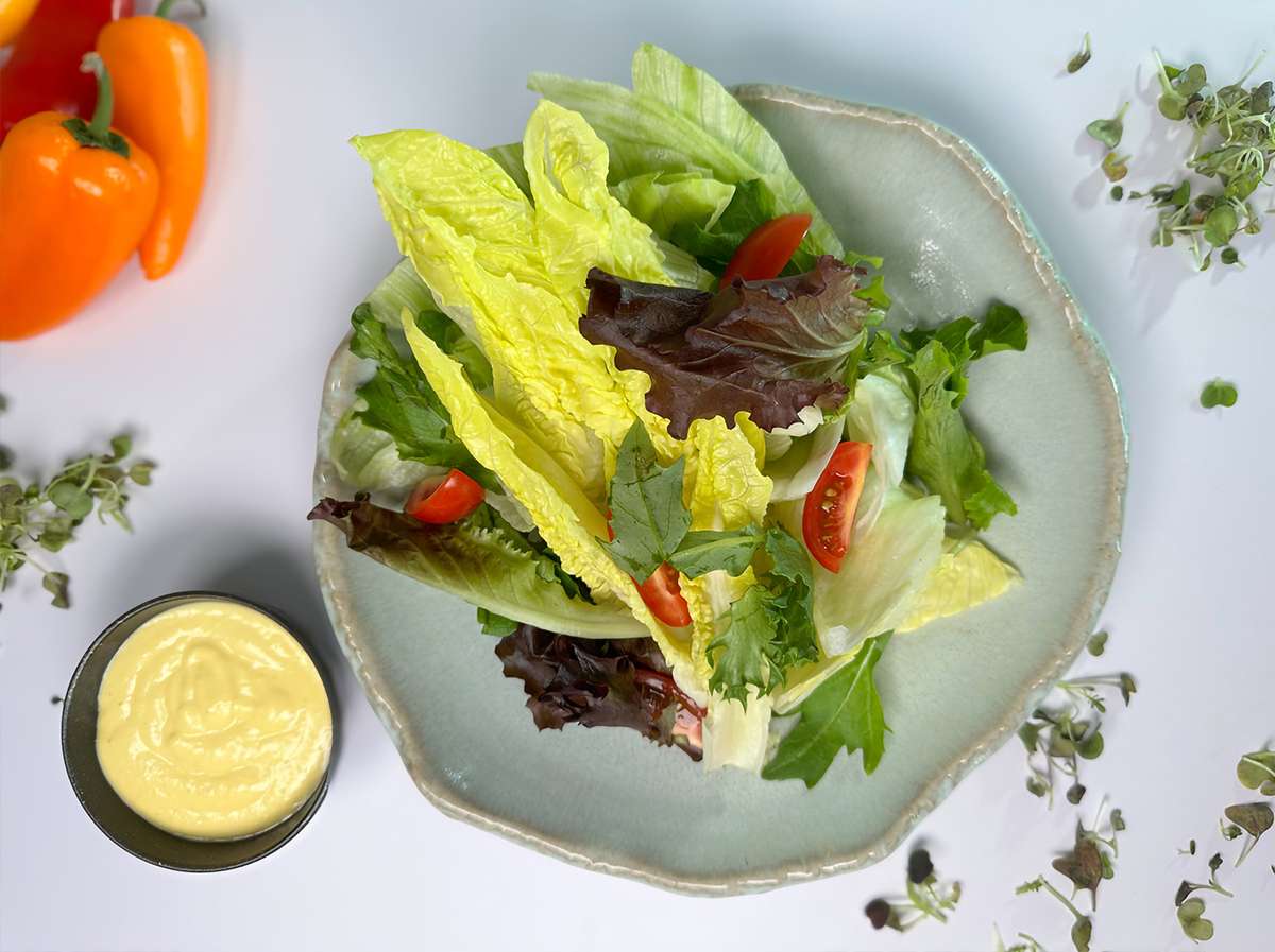 A fresh salad featuring romaine and mixed greens, cherry tomatoes, and a small bowl of creamy dressing, surrounded by colorful bell peppers and herbs on a light background.