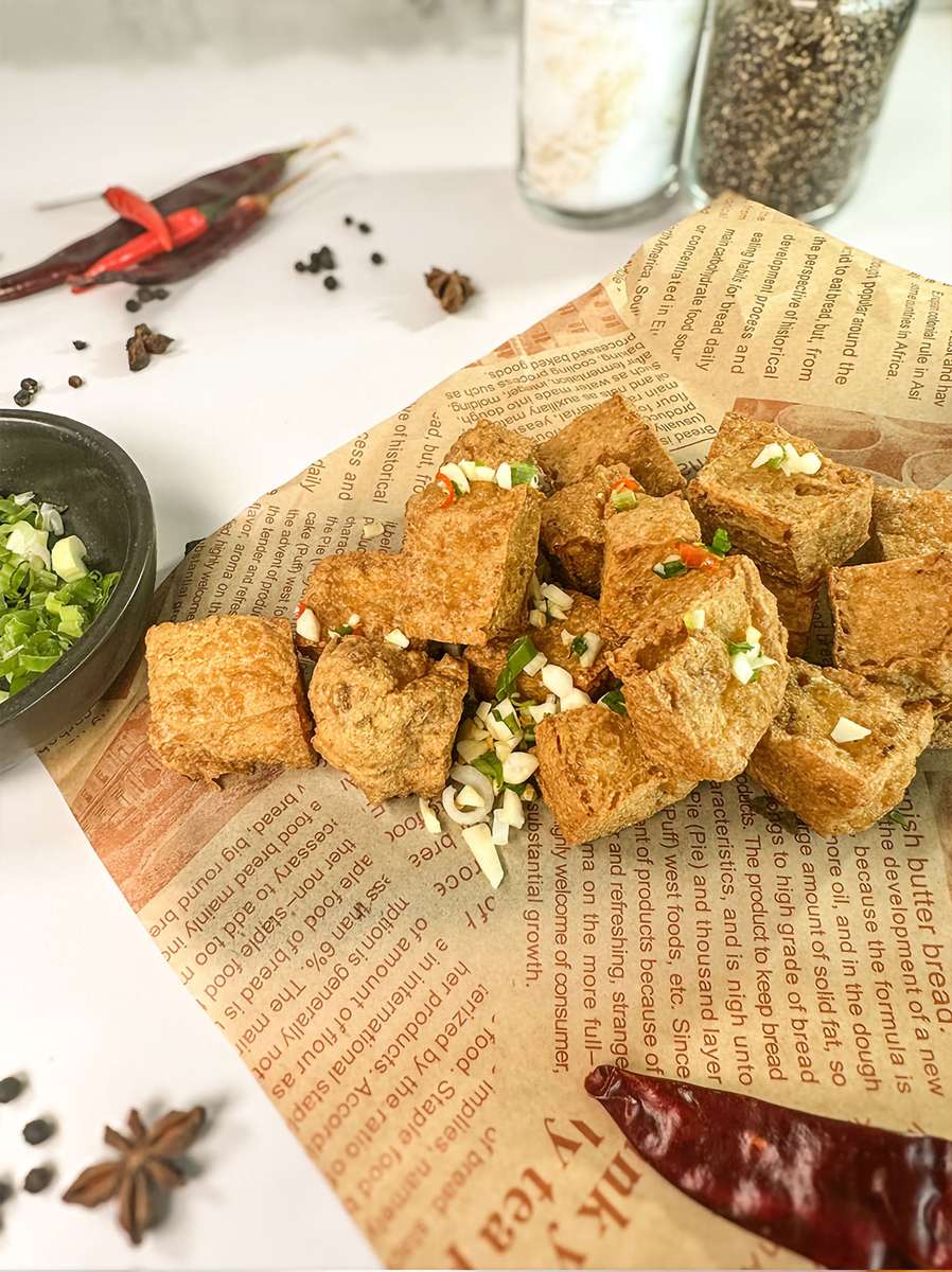 Plate of crispy fried tofu cubes garnished with chopped green onions and garlic, served on a piece of newspaper with whole spices and dried red chilies in the background.