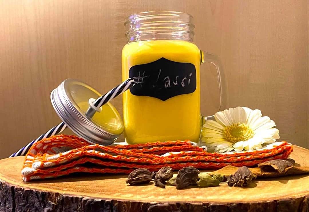 Glass mason jar of mango lassi with a striped straw, displayed on a wooden surface with whole spices, a white flower, and a colorful woven cloth.