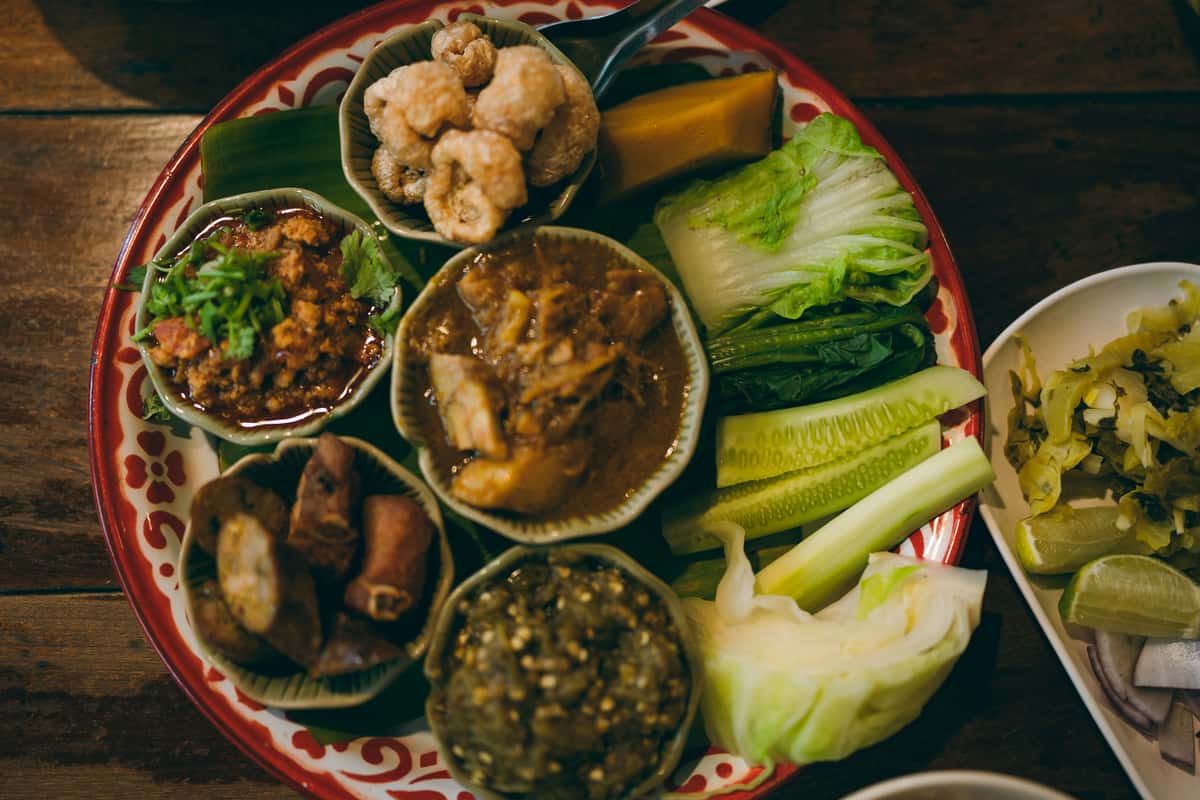 Traditional Thai platter with assorted dips, pork cracklings, curry, vegetables, cucumber, and leafy greens served on a decorative tray.
