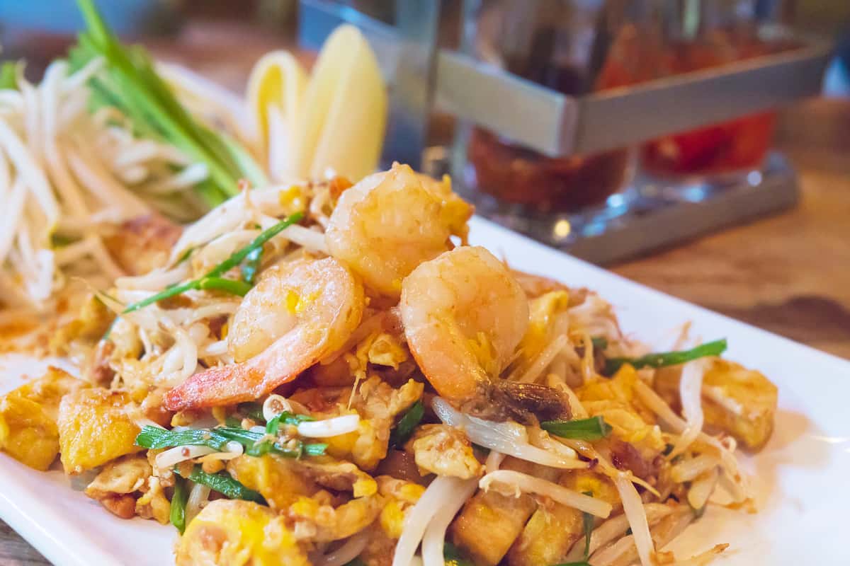 Shrimp pad thai served on a white plate with tofu, bean sprouts, scallions, and a lemon wedge in the background.
