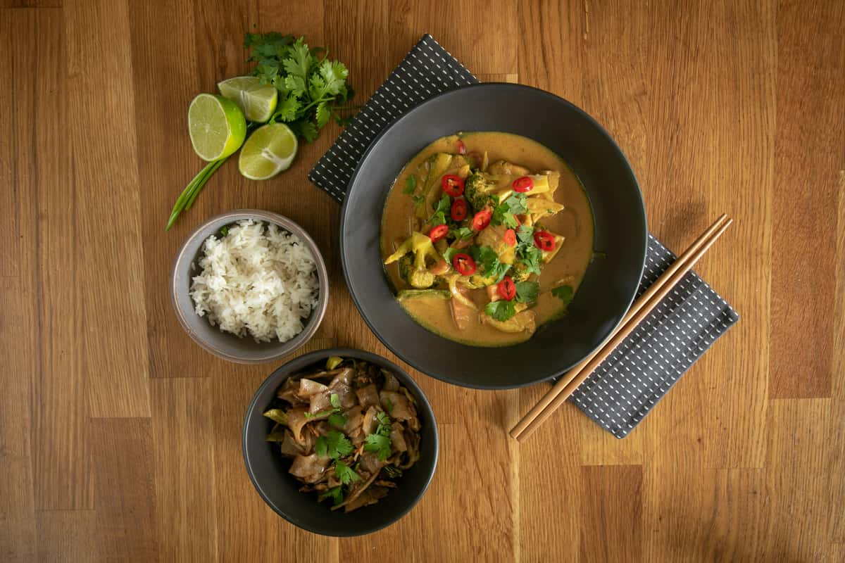 Overhead view of Thai dishes on a wooden table including curry, stir-fried noodles, jasmine rice, lime wedges, and fresh herbs.