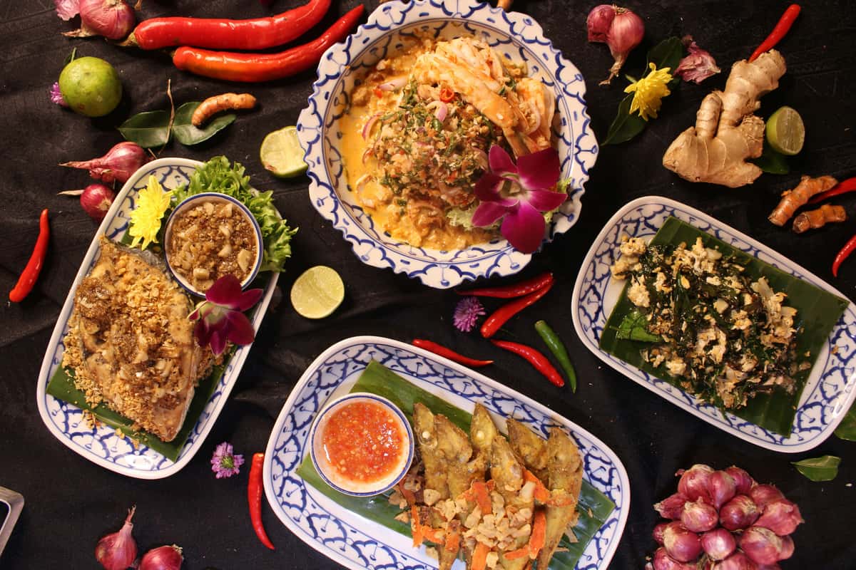 Colorful Thai dishes served in patterned bowls and plates, garnished with orchids, surrounded by fresh chilies, ginger, limes, and shallots.