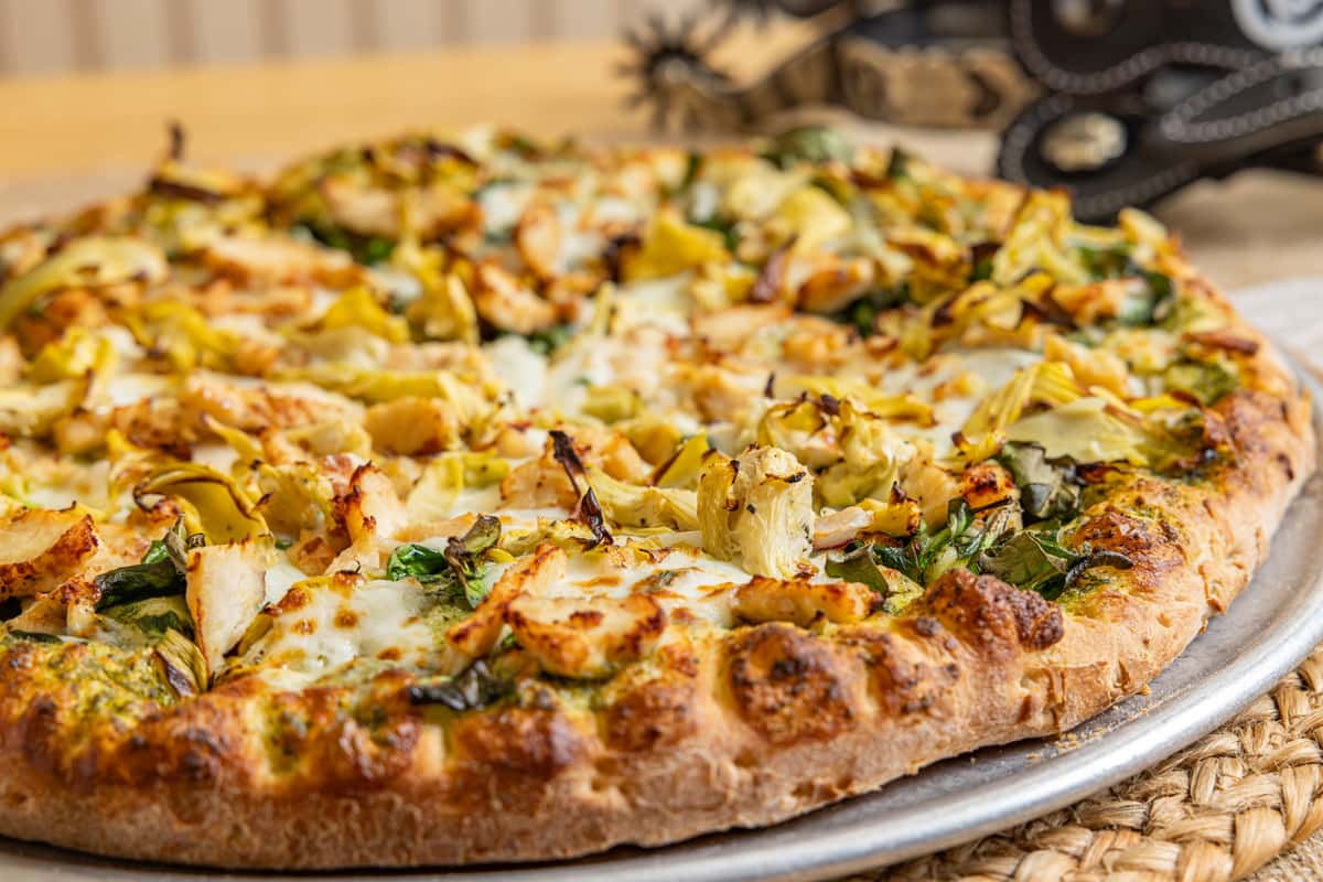 Close-up of a gourmet pesto pizza topped with grilled chicken, artichoke hearts, spinach, and melted mozzarella on a golden, hand-tossed crust.
