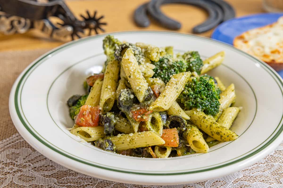 Penne pasta with broccoli, tomatoes, and black olives tossed in a creamy pesto sauce, topped with grated cheese, served in a white bowl.