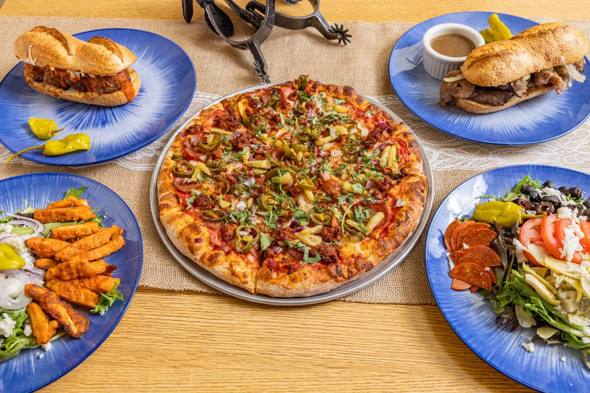 Assorted restaurant meal with a sausage and pepper pizza in the center, flanked by meatball and roast beef subs, fried chicken strips, and fresh salads on matching blue plates.