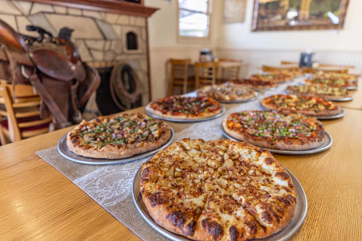 Long row of freshly baked pizzas on metal trays displayed in a rustic Western-style restaurant with a stone fireplace and saddle in the background.