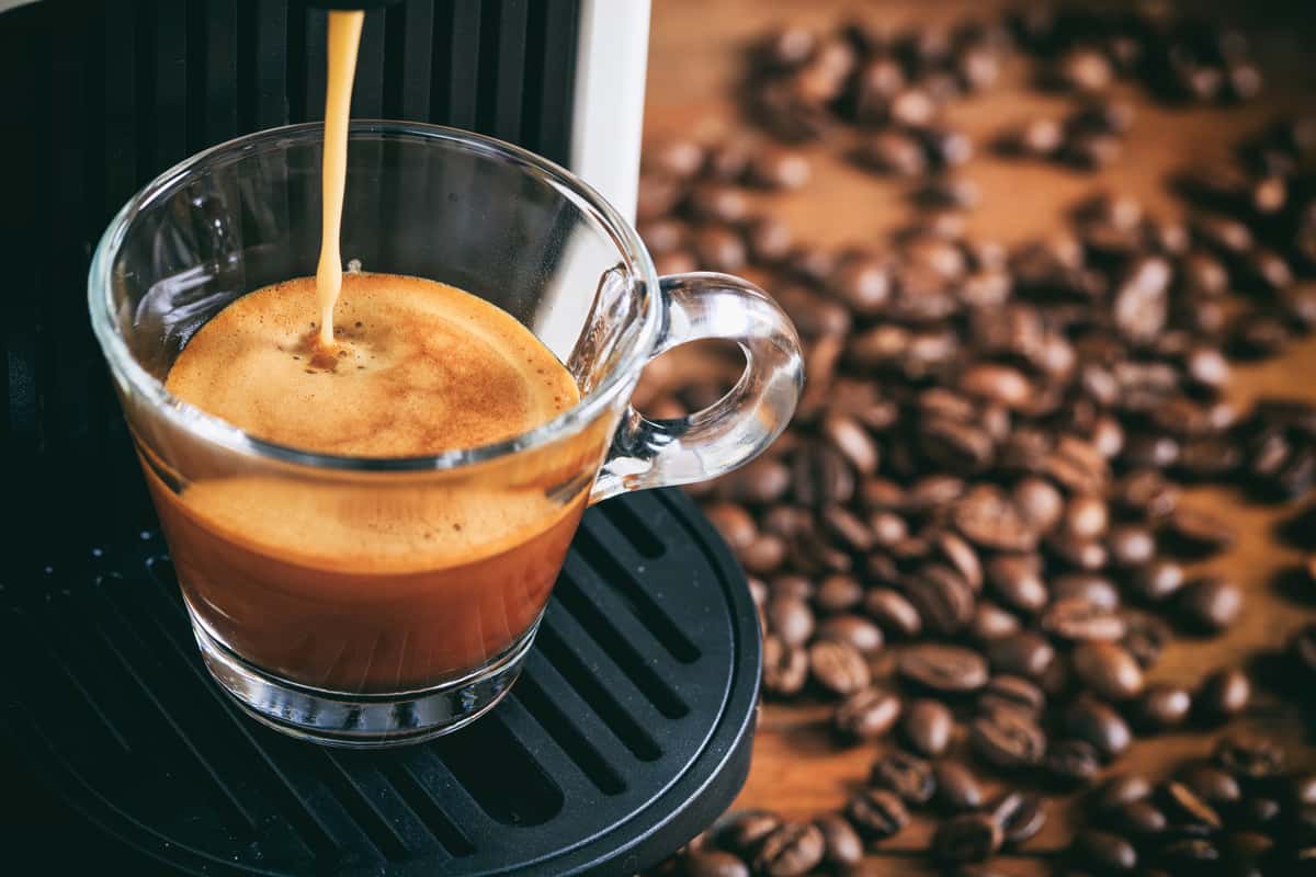 Close up of freshly brewed espresso in a glass cup beside roasted coffee beans.