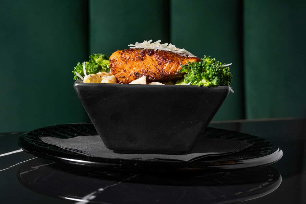 Blackened salmon filet topped with shaved Parmesan, served over creamy penne pasta and steamed broccoli in a sleek black bowl—ideal for upscale seafood dining or modern American cuisine.