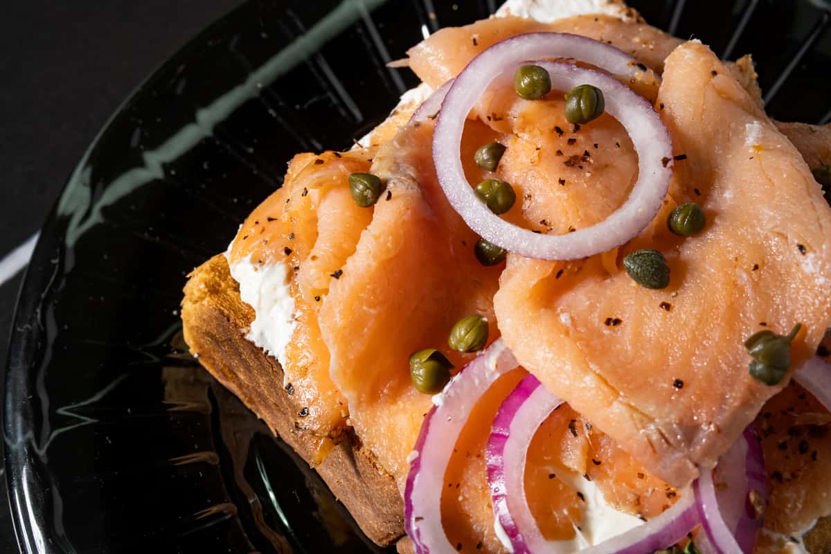 Open-faced smoked salmon toast topped with whipped cream cheese, sliced red onions, briny capers, and cracked black pepper on rustic bread—perfect for a gourmet brunch or New York-style café menu.