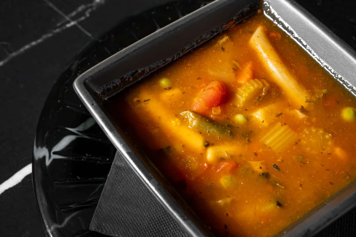 Hearty bowl of vegetable soup with penne pasta, carrots, green beans, peas, and celery in a savory tomato-based broth, served in a square black bowl—ideal for a cozy café lunch or comforting vegan appetizer.