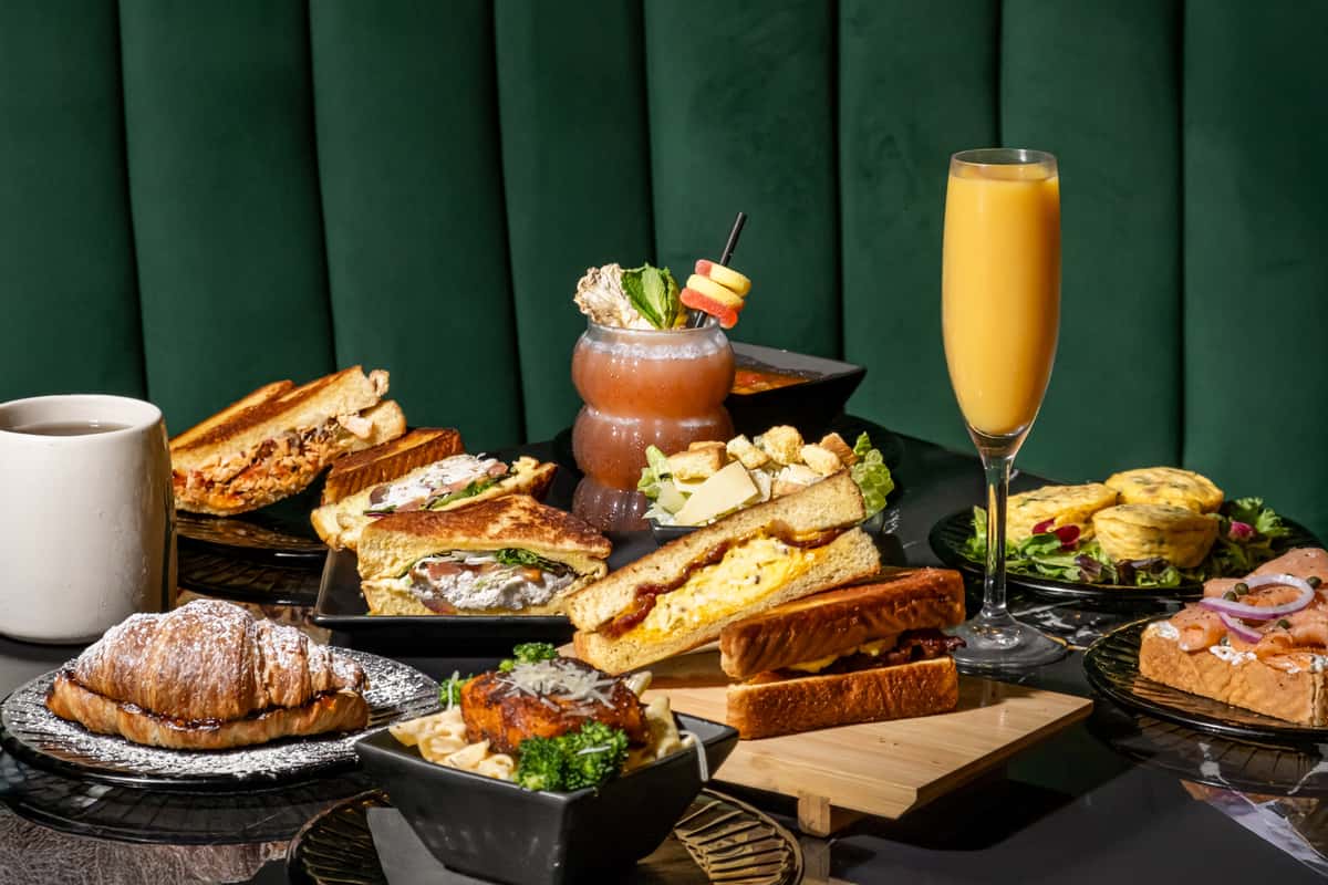 Upscale brunch spread featuring gourmet breakfast sandwiches with eggs and bacon, smoked salmon toast with capers and red onion, vegetable frittatas on greens, Caesar salad, a flaky croissant dusted with powdered sugar, creamy salmon pasta with broccoli, and beverages including coffee, a tropical cocktail, and a mimosa—set against a luxurious green velvet booth.
