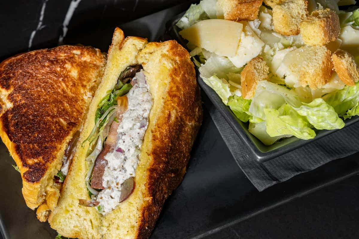 Toasted sandwich filled with creamy chicken salad, mixed greens, tomato, and red onion on golden brioche, served alongside a classic Caesar salad with romaine lettuce, shaved Parmesan, and crunchy croutons—ideal for a fresh, satisfying lunch or café favorite.