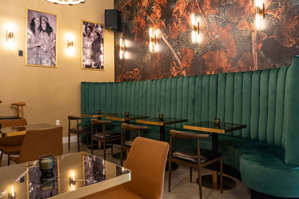 Stylish restaurant interior featuring emerald green velvet banquette seating, gold-framed portraits, moody floral accent wallpaper, and sleek black tables with candlelight—ideal for modern dining or intimate brunch settings.