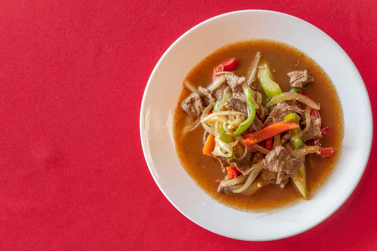Stir-fried beef with bell peppers, onions, and carrots in a savory brown sauce, served on a white plate over a vibrant red tablecloth—classic Asian-style beef stir-fry dish with colorful vegetables and rich gravy.