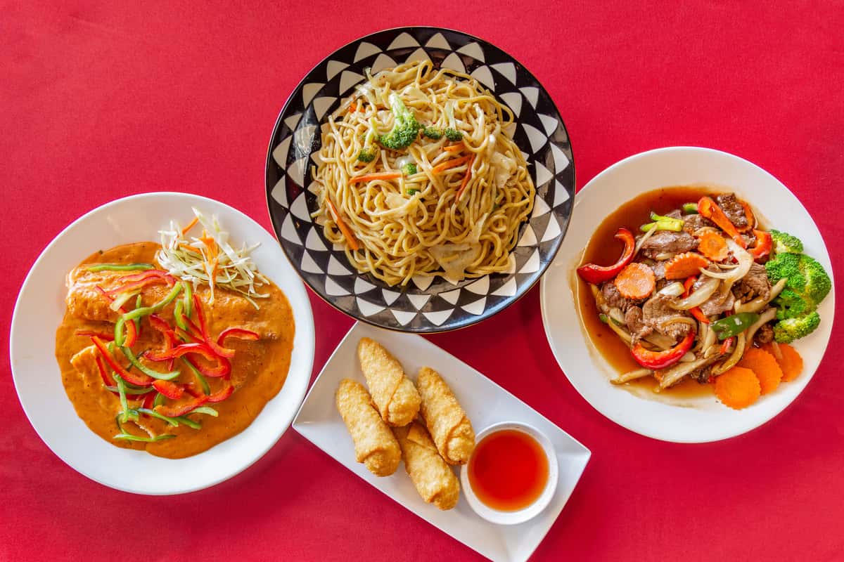Colorful Asian cuisine spread on a vibrant red table, featuring stir-fried lo mein noodles with vegetables, crispy egg rolls with sweet chili dipping sauce, Thai-style curry with bell peppers, and beef stir-fry with broccoli, carrots, and onions.