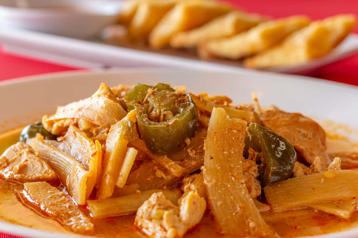 Close-up of spicy Thai bamboo shoot curry with chicken, jalapeños, and onions in a rich red chili coconut sauce, served on a white plate against a bold red tablecloth.