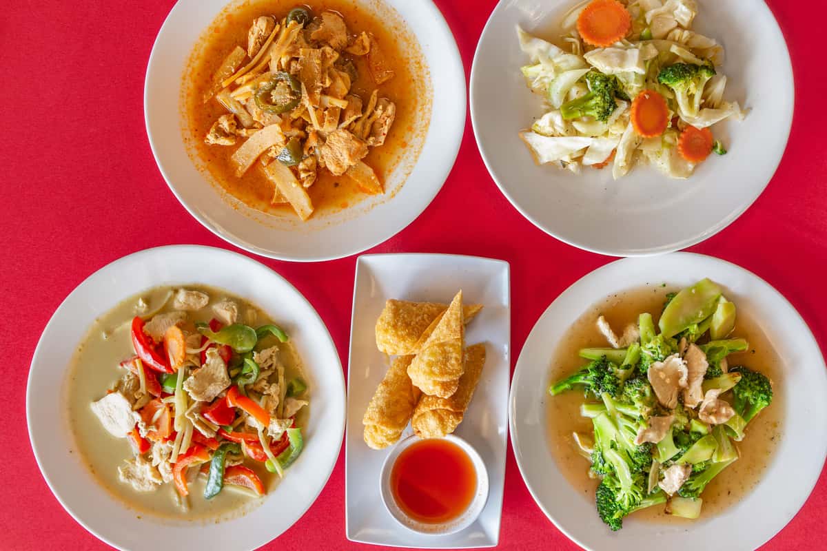 Assorted Thai dishes on white plates over a red tablecloth, featuring red curry chicken with bamboo shoots, stir-fried mixed vegetables, green curry with bell peppers, broccoli with chicken in savory brown sauce, and crispy fried wontons with sweet chili dipping sauce—colorful and flavorful Thai cuisine variety platter.