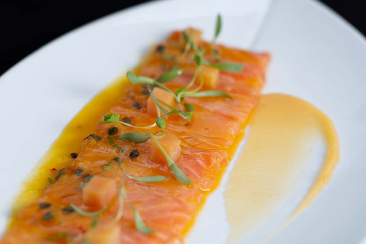 Thin slices of raw salmon arranged on a white plate with a yellow sauce and green garnish.