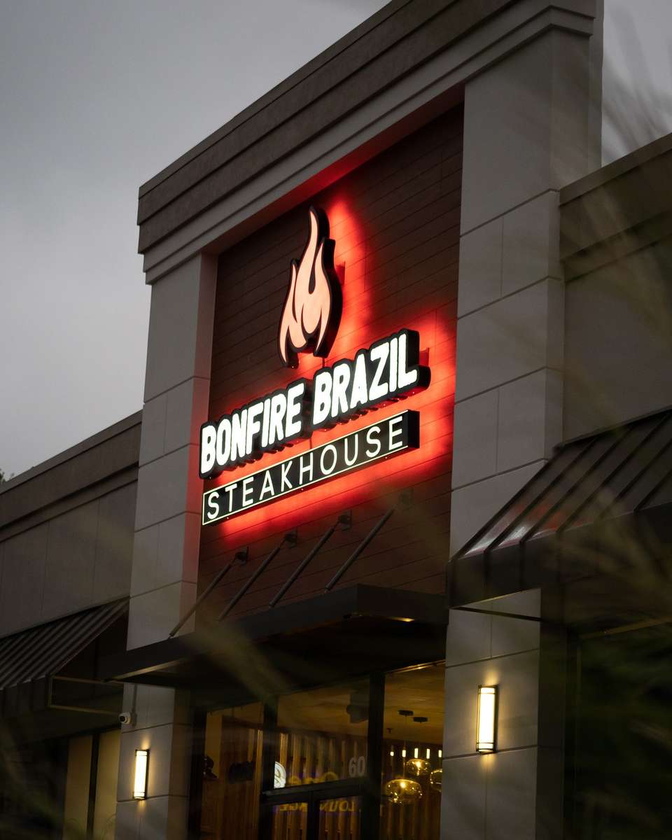 Evening exterior of Bonfire Brazil Steakhouse, featuring an illuminated flame logo and bold signage glowing in red against a wood-paneled facade. The upscale Brazilian steakhouse presents a warm, inviting ambiance with modern architectural accents and soft outdoor lighting.