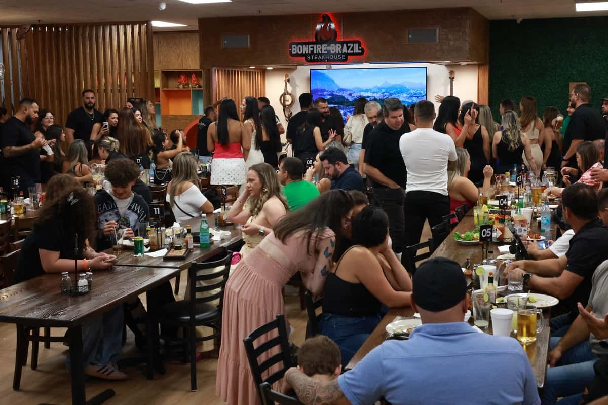 Lively evening crowd at Bonfire Brazil Steakhouse, with guests enjoying drinks and grilled meats in a vibrant, social dining atmosphere. The upscale churrascaria features long communal tables, a bustling bar scene, and a mix of families and friends celebrating together.