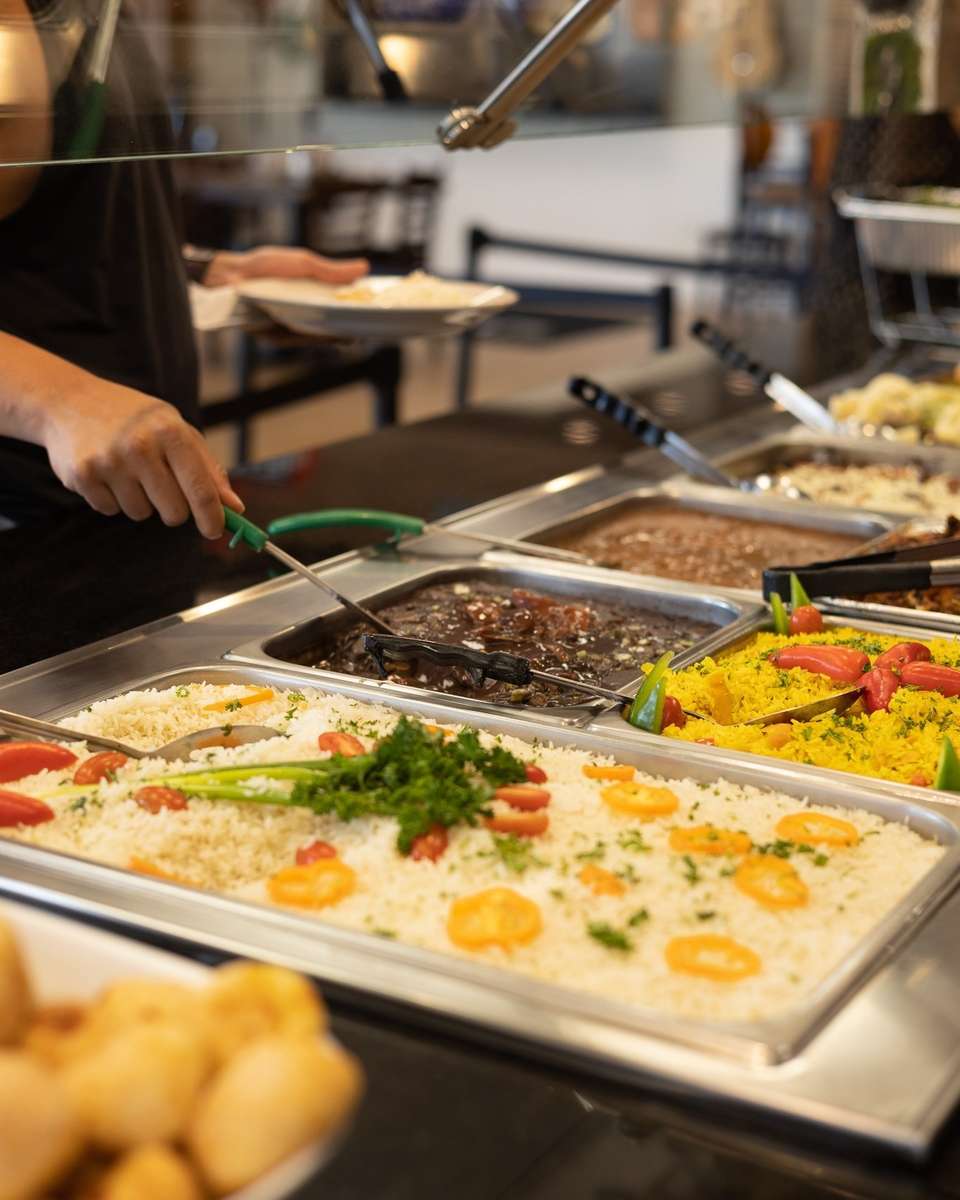 Buffet station with traditional Brazilian sides including white rice with vegetables, feijão preto (black beans), and vibrant yellow farofa, being served fresh during lunch or dinner service.