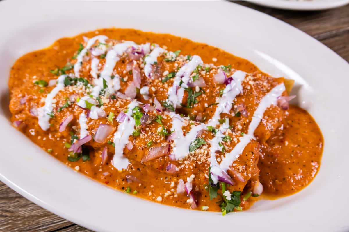 Creamy shrimp enchiladas topped with rich chipotle sauce, sour cream, diced red onions, cilantro, and crumbled queso fresco on a white plate.