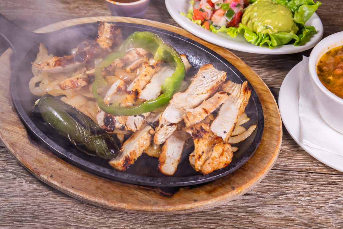 Sizzling grilled chicken fajitas served on a hot cast-iron skillet with sautéed onions, green bell peppers, roasted jalapeño, fresh guacamole salad, and a side of charro beans.