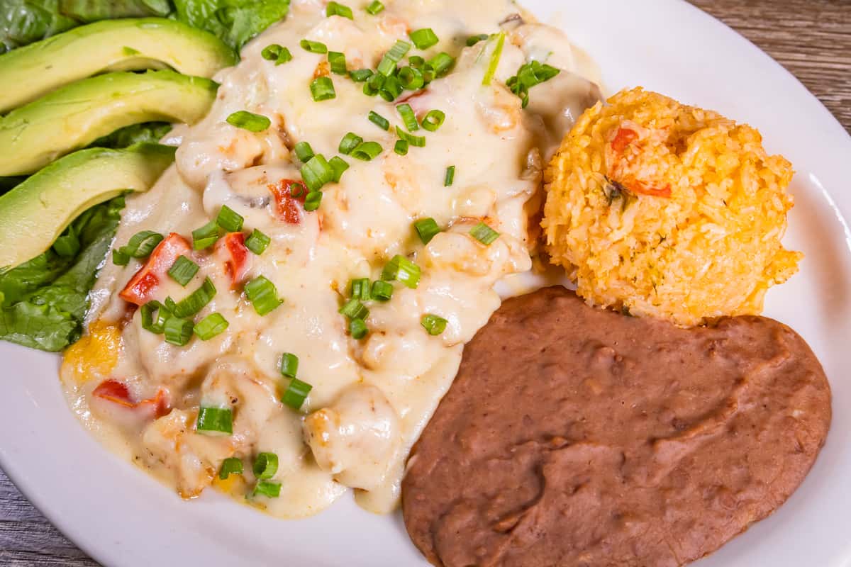Grilled chicken breast topped with creamy shrimp sauce and melted cheese, served with avocado slices, Mexican rice, refried beans, and crisp lettuce on a white plate.