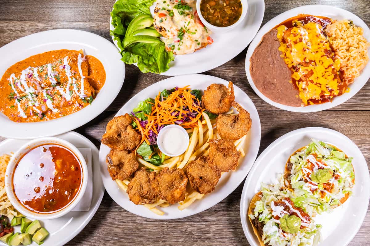 Assorted Mexican dishes including fried butterfly shrimp, enchiladas, tostadas, grilled seafood, hearty soup, and rice and beans served on a restaurant table.