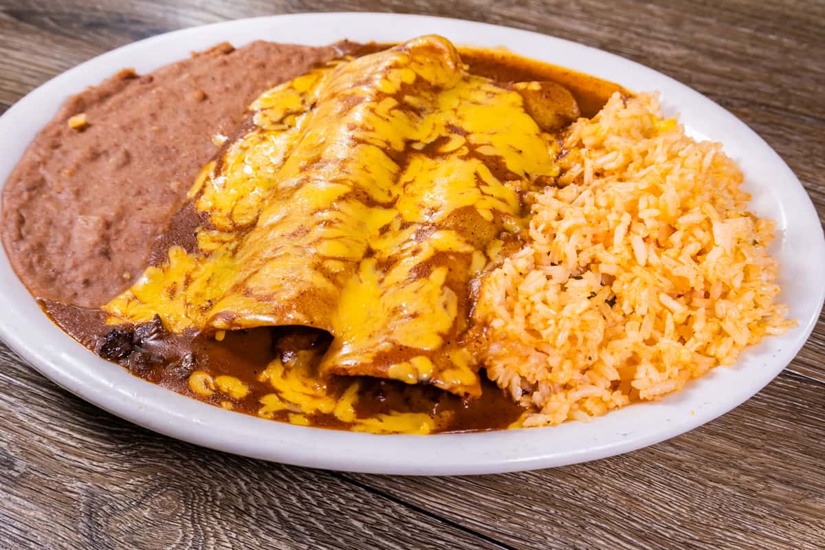 Cheese-covered beef enchiladas topped with savory chili gravy, served with Mexican rice and refried beans on a white plate.