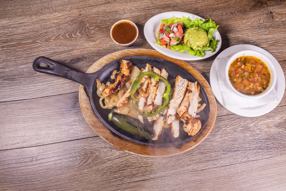 Sizzling grilled chicken fajitas served on a cast-iron skillet with sautéed onions, green bell peppers, roasted jalapeño, fresh guacamole, pico de gallo, salsa, and a side of charro beans.