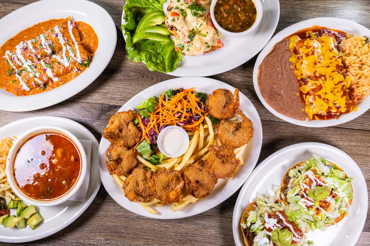 Assorted Mexican favorites including fried butterfly shrimp, cheese enchiladas, tostadas, grilled seafood, hearty soup, and traditional sides served on a restaurant table.