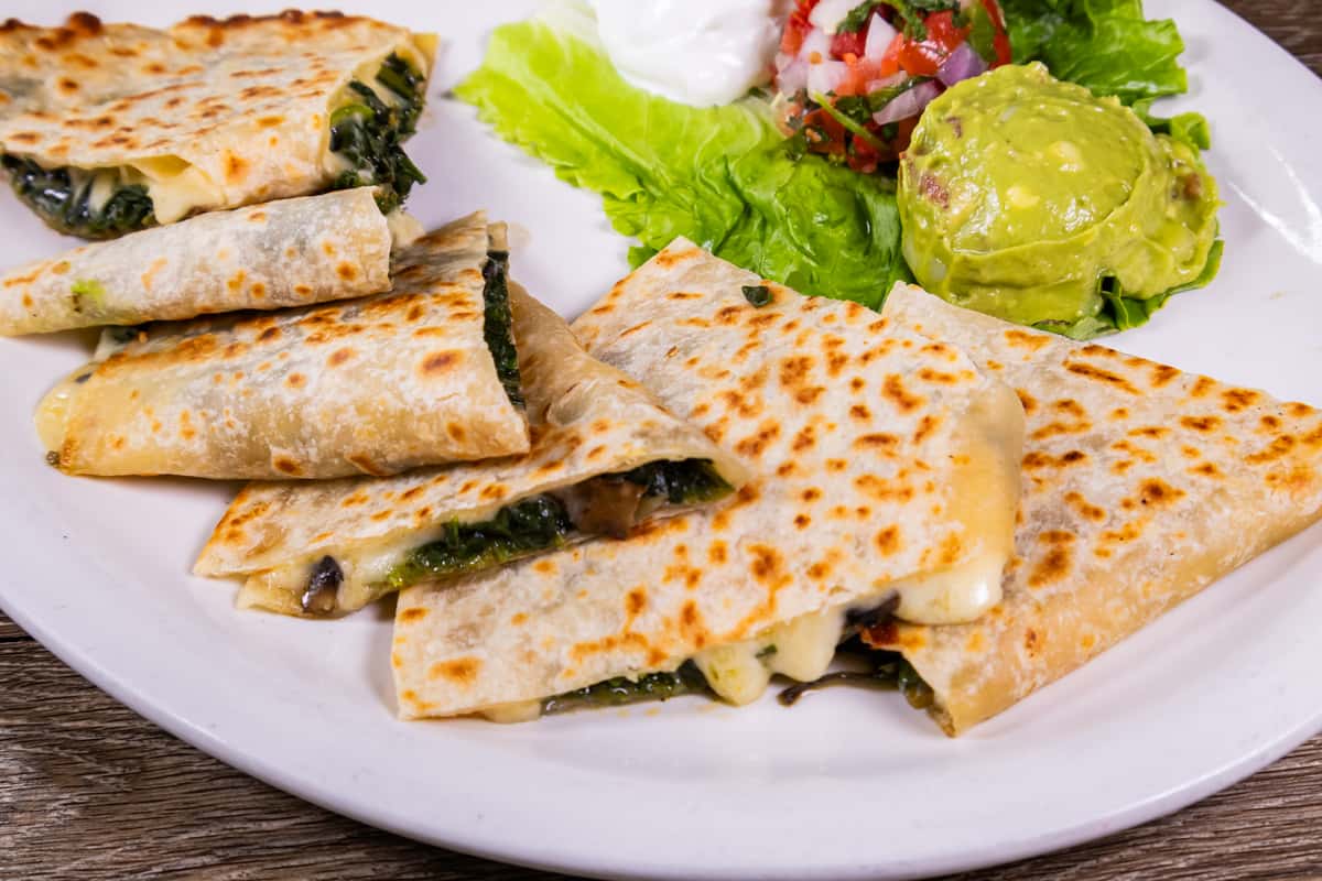 Grilled spinach and cheese quesadillas served with fresh guacamole, pico de gallo, sour cream, and crisp lettuce on a white plate.