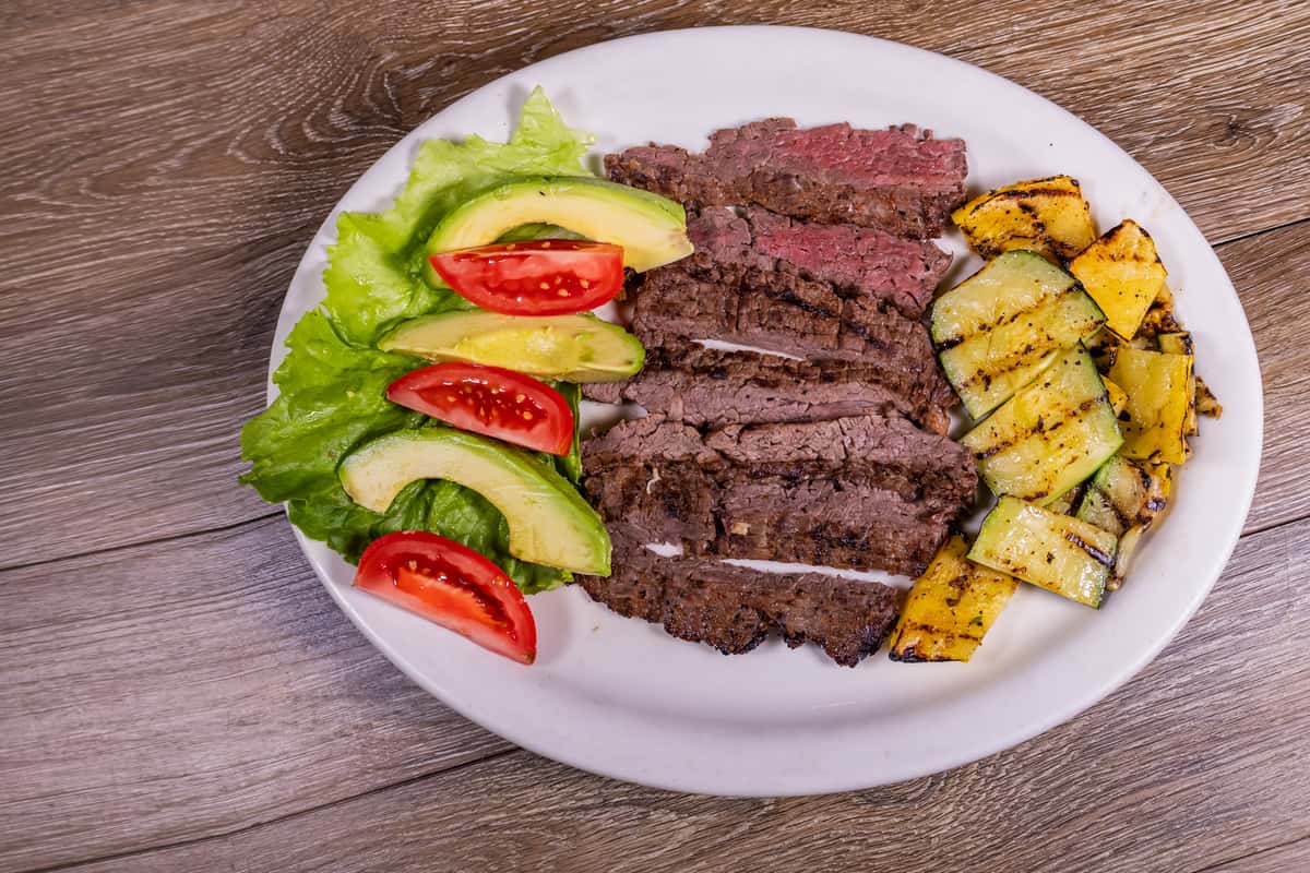 Grilled sliced steak served with fresh avocado, tomato wedges, crisp lettuce, and grilled zucchini and squash on a white plate.
